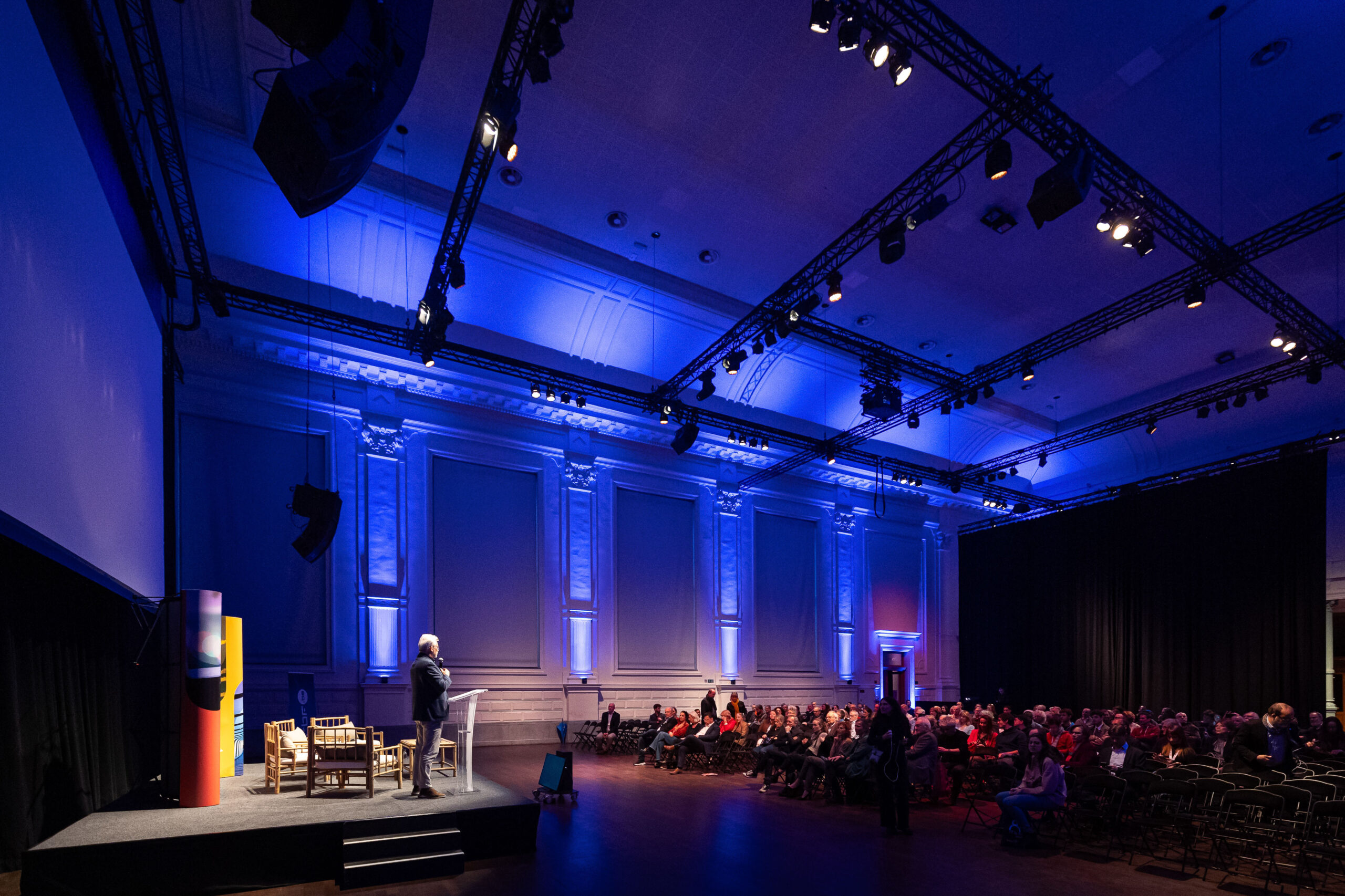 Photographe événement capturant un conférencier sur scène devant un auditoire en salle de conférence éclairée en bleu, avec projecteur et installations scéniques.