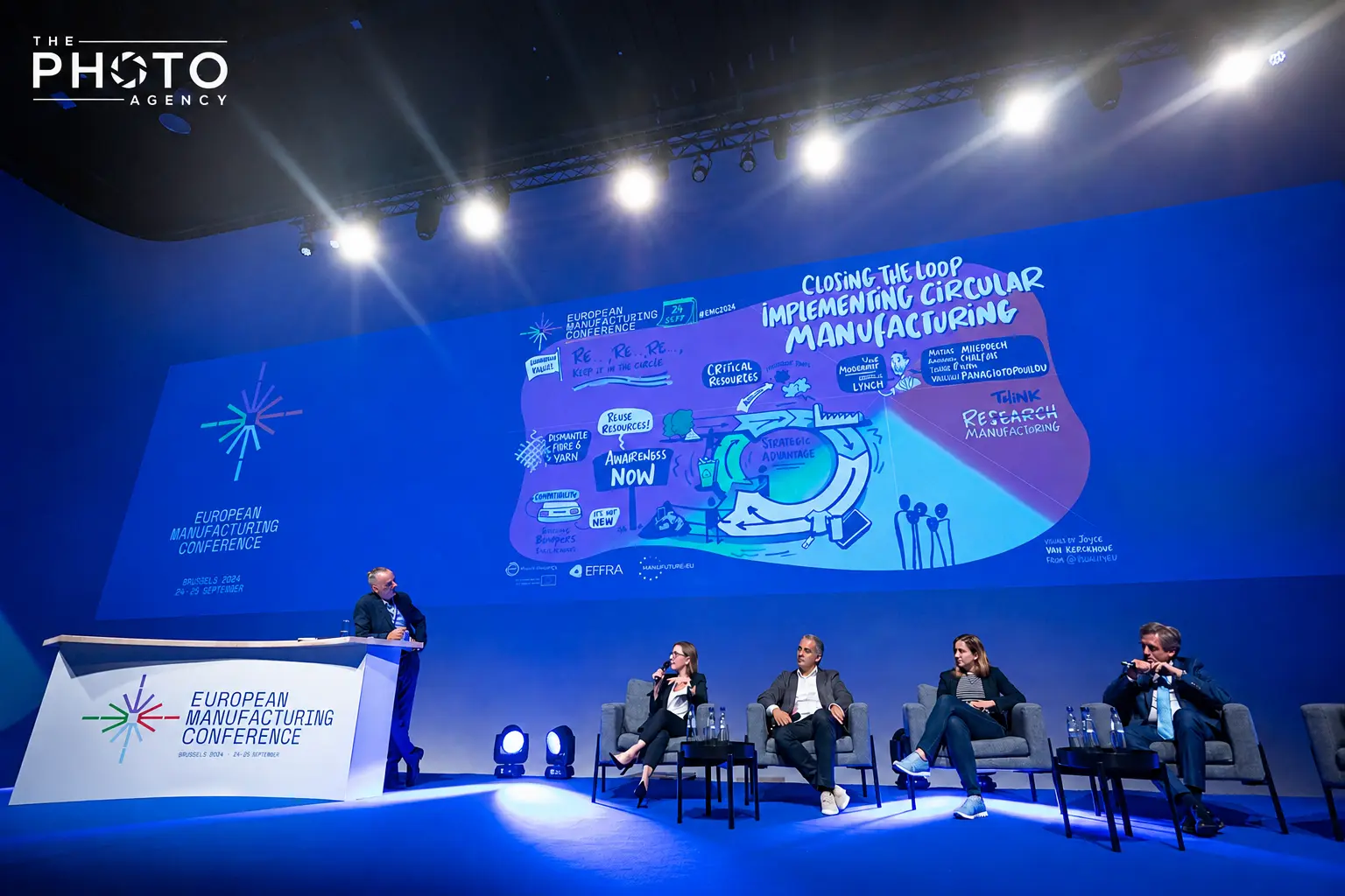 Conférence européenne sur la fabrication, avec un podium et panneaux bleus, orateurs assis sur scène et une grande présentation graphique illuminée.
