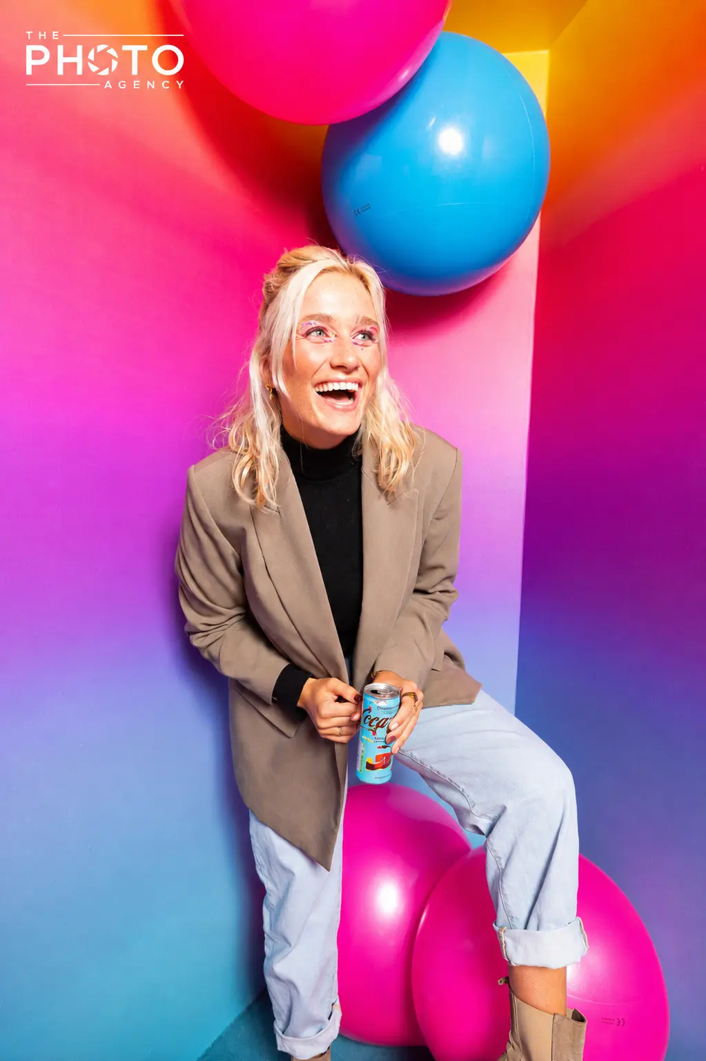 Femme blonde souriante dans un décor coloré avec ballons, porte un blazer beige et un col roulé noir, assise près de ballons roses et bleus, en train de boire dans une canette.