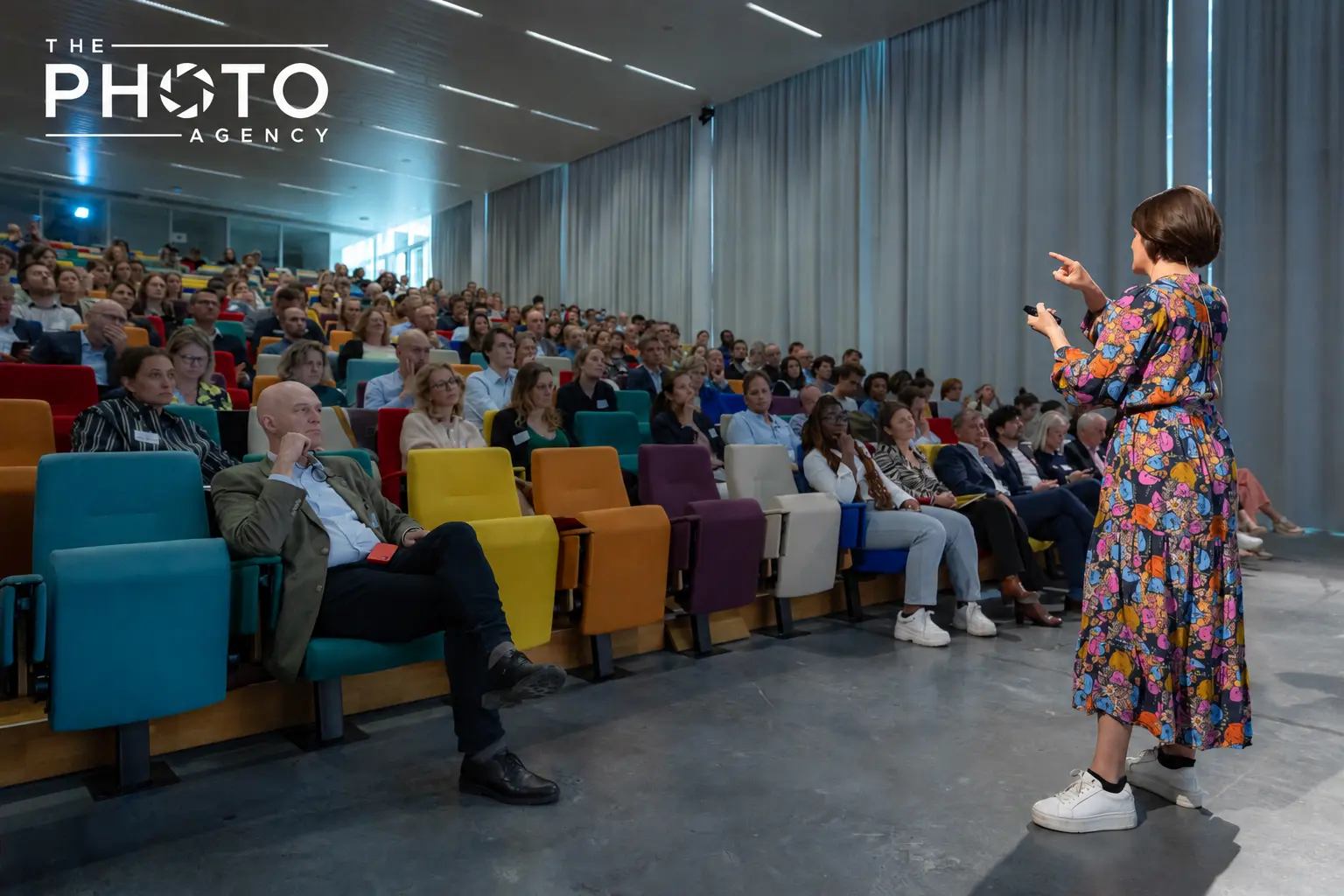 Conférencière en robe à motifs colorés s’adressant à une audience captivée dans une salle de conférence multi-seats colorés, The Photo Agency.