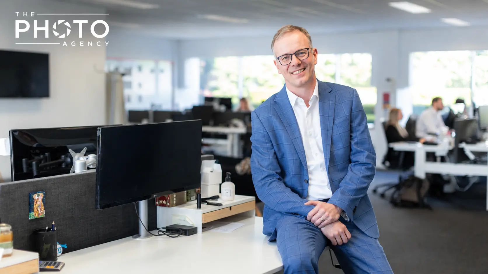 Portrait d’un homme d’affaires en veste bleue dans un open space moderne, assis près d’un bureau avec écran et accessoires. The Photo Agency.